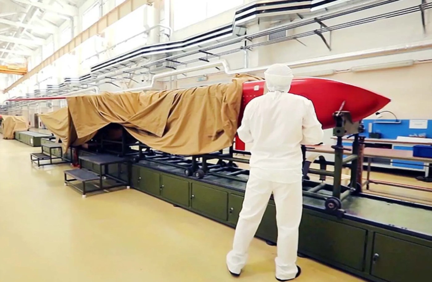 Worker in white overalls beside a tarpaulin-covered missile with a red nose section in a factory; a video frame of unconfirmed location and date