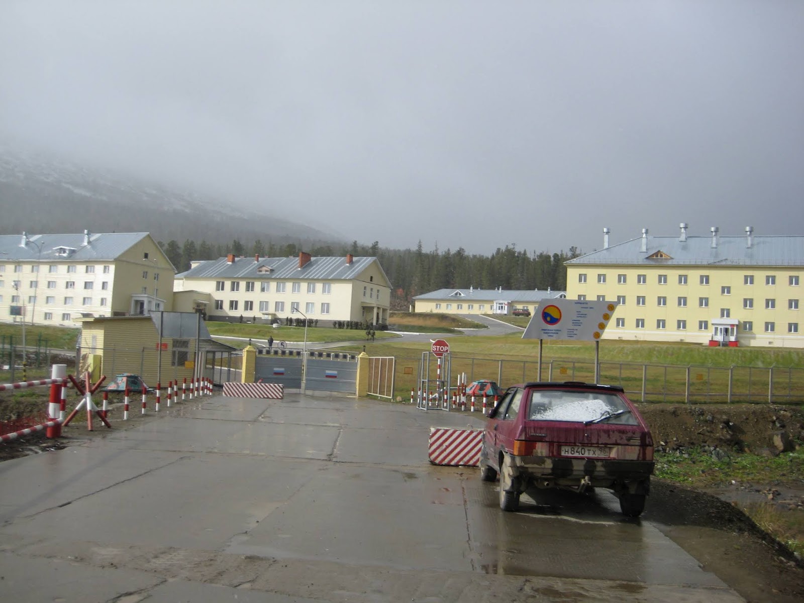 Gate, red-and-white barriers, STOP sign, red car and yellow two-storey buildings of a military base under low cloud