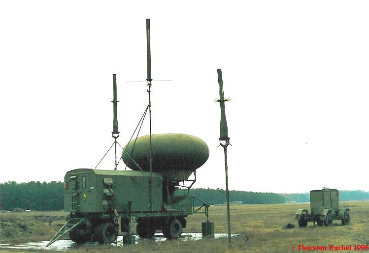 Radome trailer with three masts and a second small trailer beside a pine forest